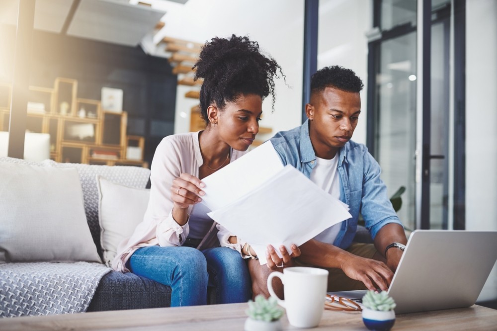 A couple sitting together on a couch, intently examining papers and working on a laptop.