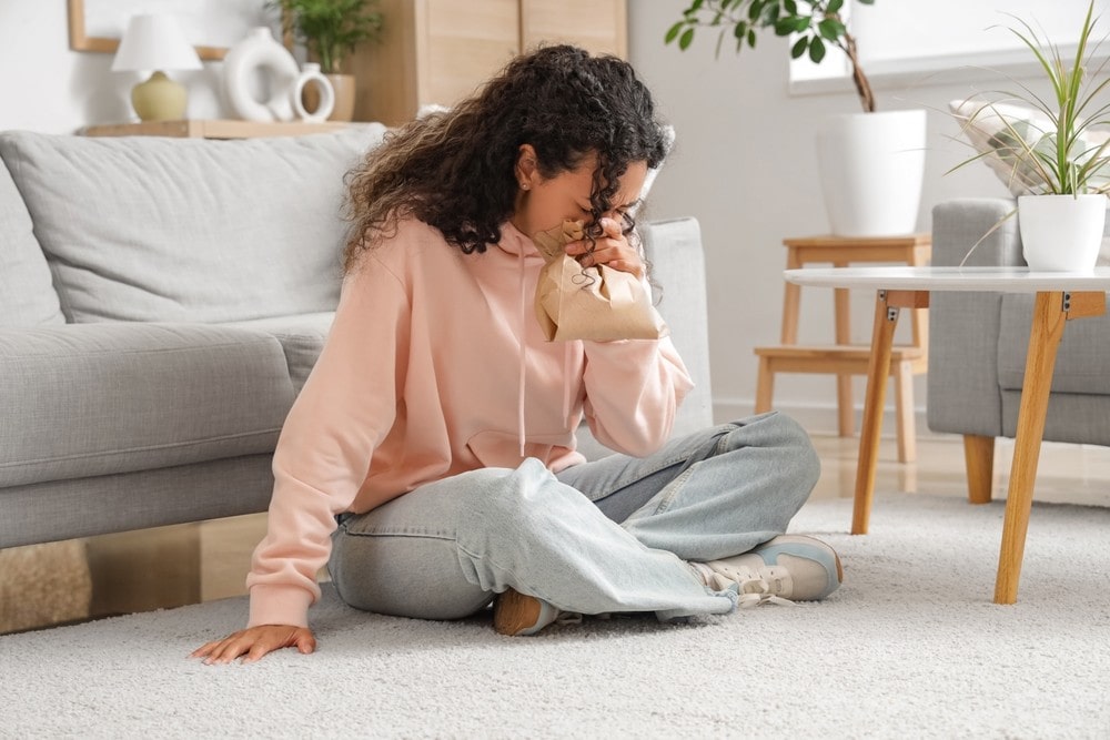 A young woman sits on the floor, breathing into a paper bag.