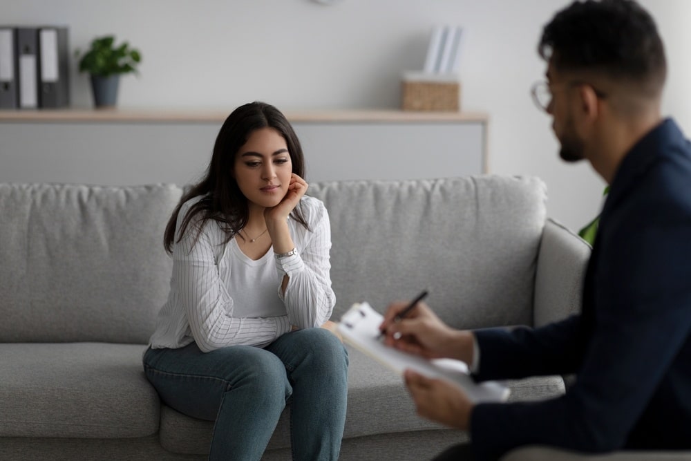 A woman sits on a couch, looking sad and thoughtful, while a man sits across from her, taking notes.