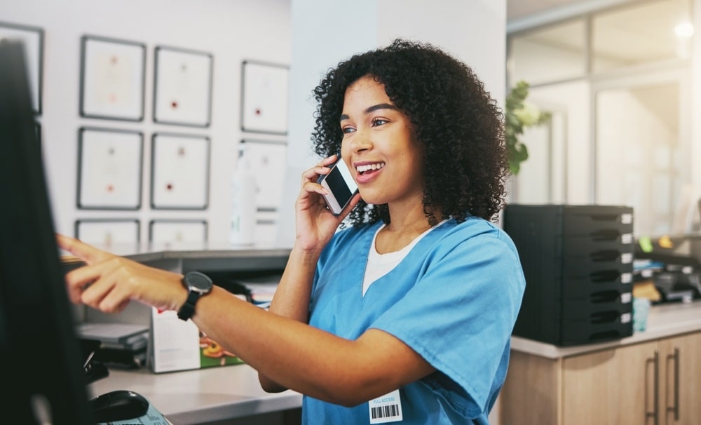 A smiling woman in blue scrubs is on her phone while working at a computer.
