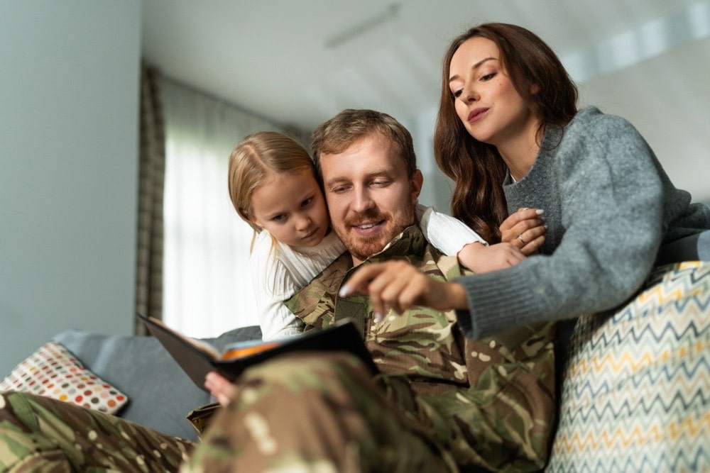 A man in military uniform sitting on a couch with a woman and a young girl, all engaged in looking at a book together.